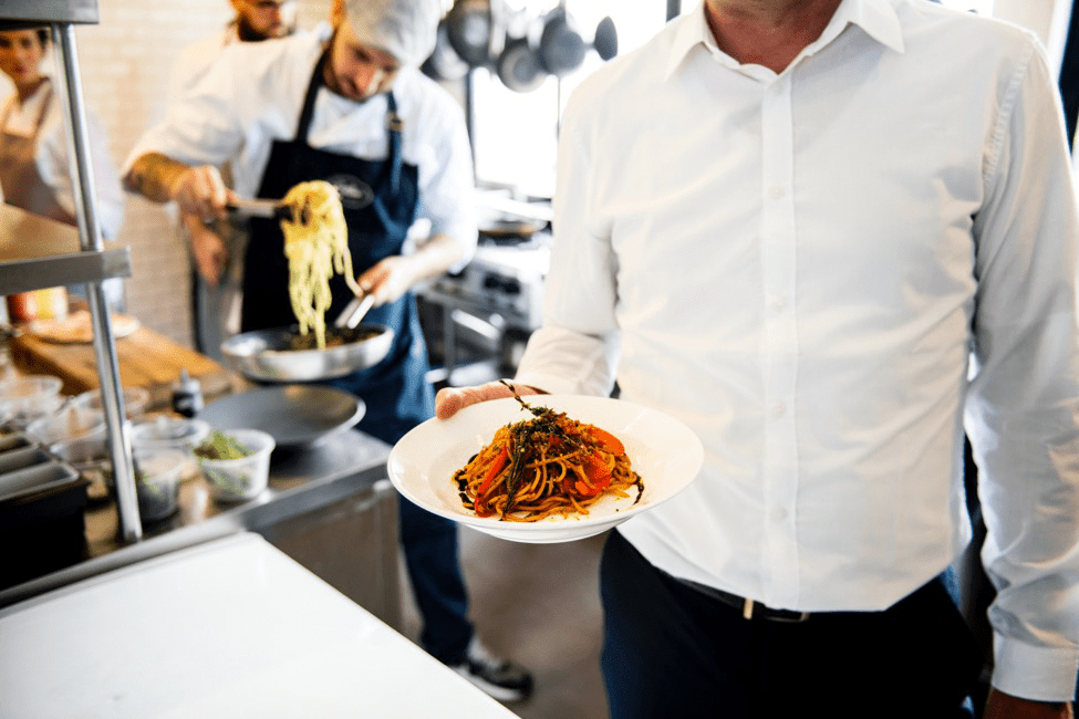  a waiter holding a pasta dish while a cook prepares another dish in the background