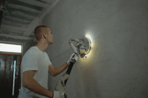 A male worker sanding a wall by using a sander with a light on it.