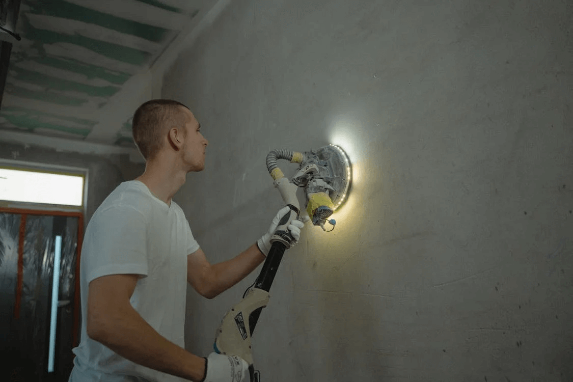 A male worker sanding a wall by using a sander with a light on it.