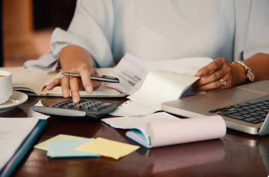 A person using a calculator, paper, and pen, with a laptop pushed to the side of the desk.