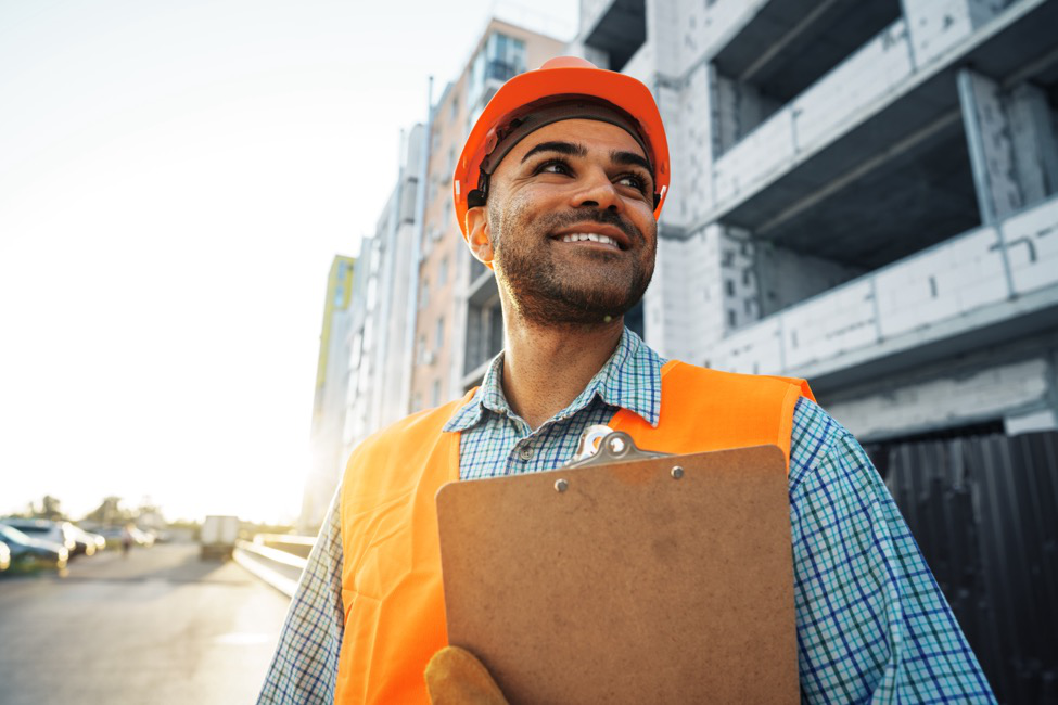 A man wearing PPE stands holding a clipboard at a construction site.
