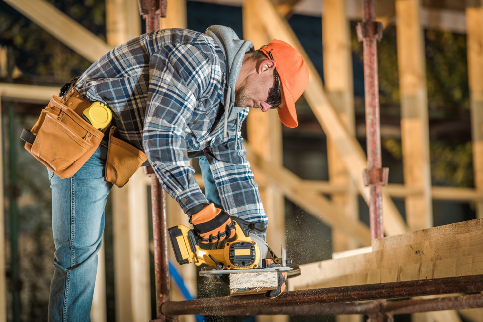 A skilled labor carpenter wearing a hard hat, safety glasses, and a tool belt saws a wooden plank.