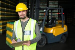 A warehouse supervisor in a yellow safety vest and hard hat holding a clipboard with a forklift in the background