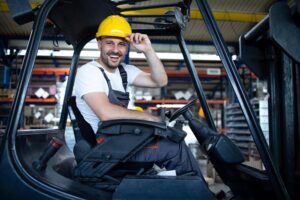 A smiling forklift operator wearing a yellow hard hat and safety gear while operating machinery in a busy warehouse.