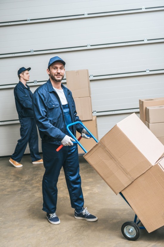 A general labor worker pushes two boxes on a dolly in an industrial warehouse.