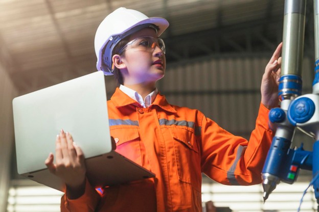 A female technician wearing an orange jumpsuit, safety glasses, and a white hard hat holding a laptop while monitoring industrial machinery.