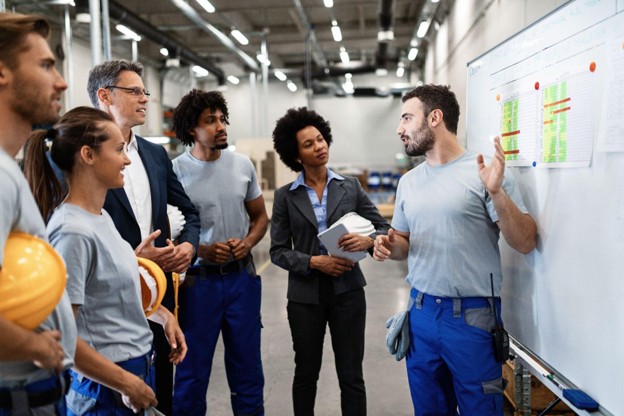 A diverse group of factory staff and corporate managers standing around a whiteboard in an industrial plant to plan shift schedules.
