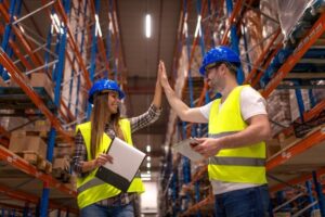 Two smiling logistics workers wearing blue hard hats and yellow safety vests high-fiving in a tall warehouse storage aisle.